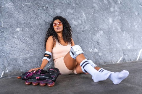 Stylish urban latin woman sitting with inline skates leaning against grey wall in the street