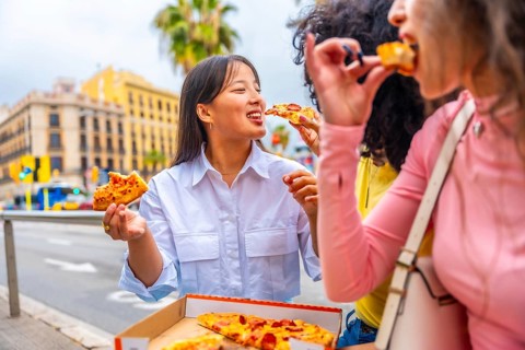 Three multicultural female friends sharing and eating pizza sitting outside