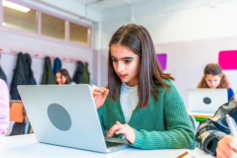 Schoolgirl concentrating on her laptop during a lesson, surrounded by classmates in a modern