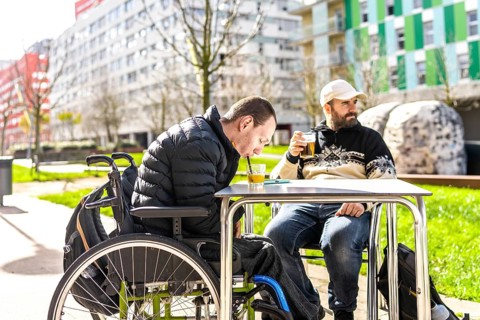 Two men enjoy drinks at an outdoor cafe, one using a wheelchair, demonstrating inclusivity and