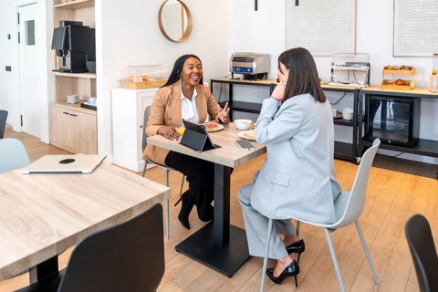 Two businesswomen enjoying breakfast and discussing work during a business trip, utilizing a tablet