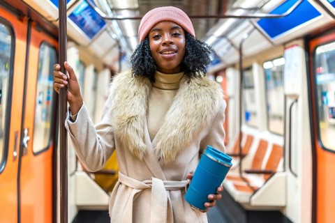 Smiling young black woman wearing winter coat and pink wool hat holding reusable coffee cup while