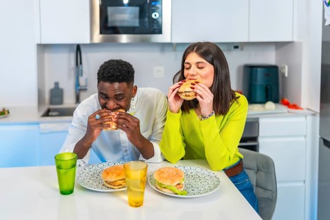 Young multiethnic couple eating homemade burgers in their modern kitchen, enjoying a casual meal