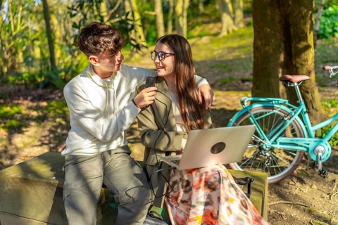 Two students are working on a laptop in a park, charging it with a power bank while sitting on a