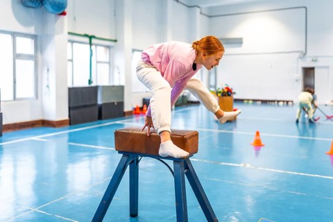 Girl practicing gymnastics, balancing on pommel horse in school gym, showing determination and