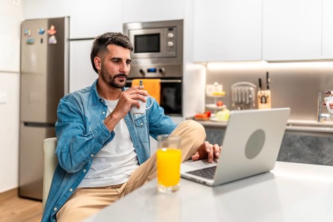 Man savoring a cup of coffee while working remotely on a laptop in a stylish, modern kitchen,