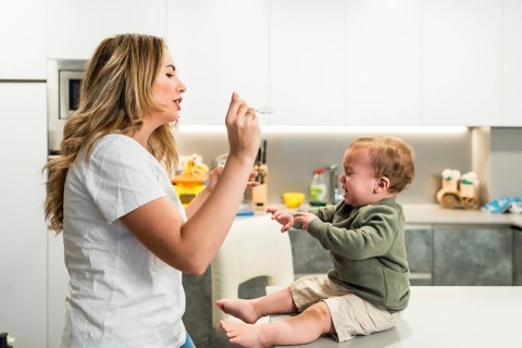 Young mother struggling to feed her crying baby boy while sitting on the kitchen counter at home,