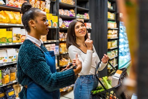 Saleswoman using tablet, advising customer holding shopping basket, choosing products on shelves in