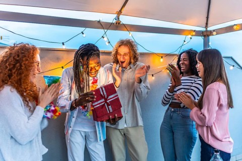 Happy young african man opening birthday gift with multi-ethnic friends during roof party