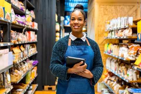 Confident saleswoman in an apron, smiling while holding a digital tablet and working among shelves