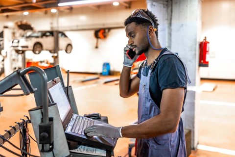 Young mechanic using a laptop and talking on the phone while working on a car in a professional