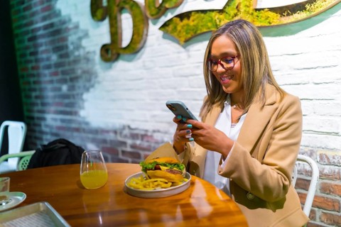 Beautiful latin woman wearing business casual clothes enjoying tasty burger and texting on the
