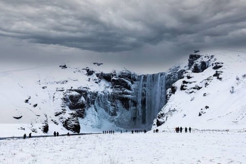 Tourists at Skogafoss waterfall, snowy and icy rock face, dark clouds above, Sudurland, Iceland,