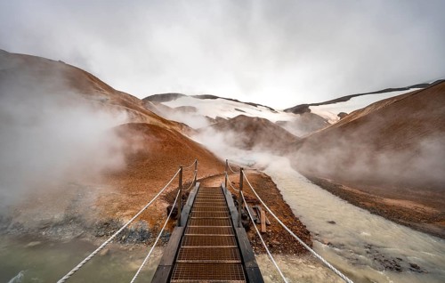 Bridge over steaming stream between colourful rhyolite mountains with snowfields, Hveradalir