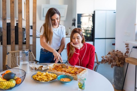 Two women are slicing and sharing a rectangular pizza, nachos with sauce and tacos at home in their