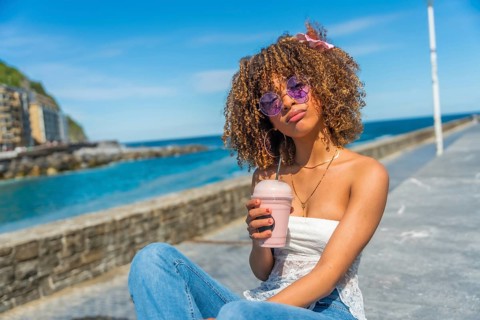 Cool latin young woman with curly hair in summer clothes drinking a shake outdoors next to the sea