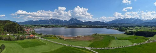 Expansive landscape with a view of Lake Hopfensee and mountains, surrounded by green meadows and a