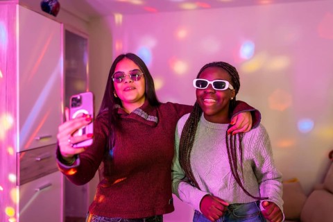 Two happy teenage girls with sunglasses taking a selfie with smartphone at a home party with disco