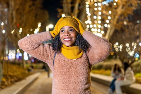 Smiling young woman wearing winter clothes adjusting her yellow wool cap under christmas lights in