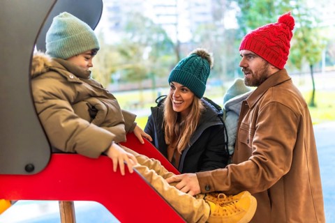 Smiling parents watching their son going down a slide at the playground on a cold winter day