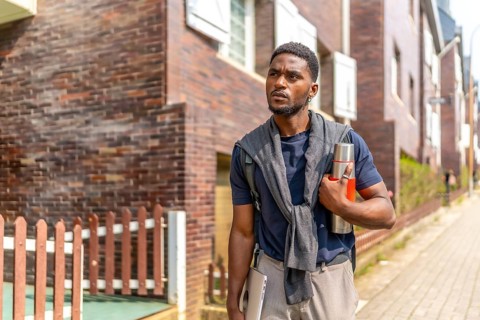 Young african entrepreneur walking the street with reusable coffee thermos and laptop