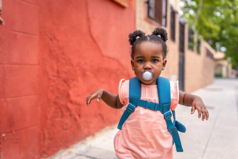 Cute baby african girl running along a street using pacifier