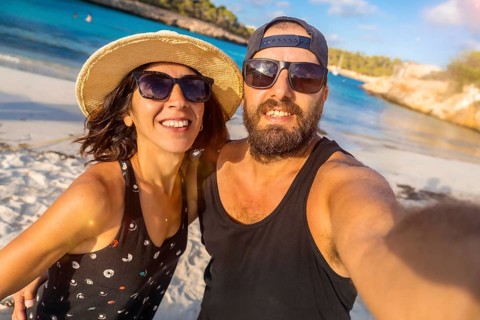 A man and woman are smiling for the camera on a beach. The man is wearing a black tank top and