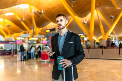 Businessman holding a reusable coffee cup and smartphone while waiting for his flight at the
