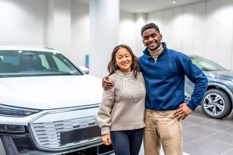 Happy multiethnic couple hugging, buying new electric car at dealership, embracing modern eco