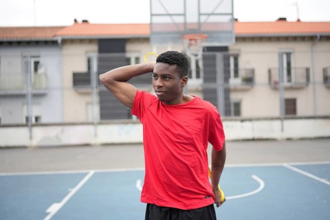 African boy stretching using rubber elastic band alone before playing basketball outdoors