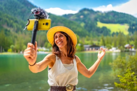 Smiling woman wearing straw hat vlogging with smartphone and external microphone at lake dobbiaco