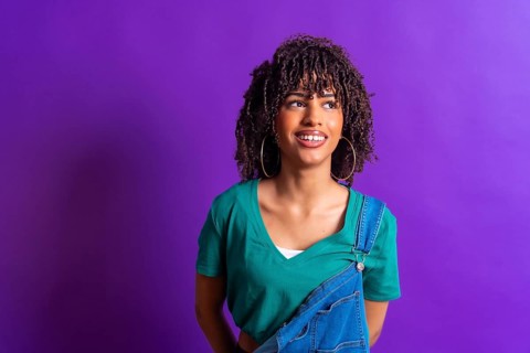 Young woman with curly hair, wearing a green t shirt and denim overalls, smiles while looking up
