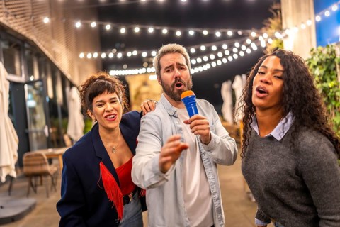 Man and two multiracial women singing karaoke having fun at night party in a terrace of a bar
