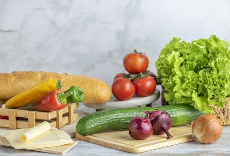 Colourful peppers, tomatoes, cucumber, lettuce and slices of cheese next to a baguette on a table,
