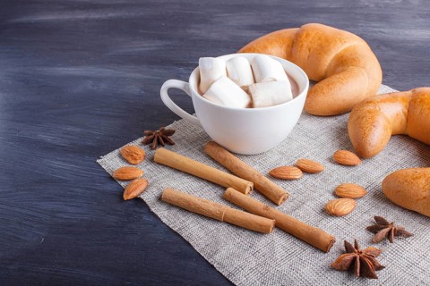 A cup of hot chocolate with marshmallow, croissants and spices on linen napkin and black wooden