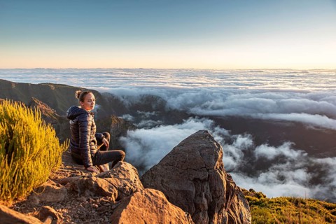 A woman sits on a rocky mountainside and calmly enjoys the wide view over the sea of clouds, Pico