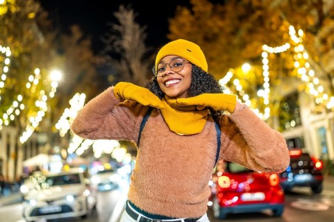 Smiling young woman wearing yellow winter clothes enjoying christmas lights in a city street at