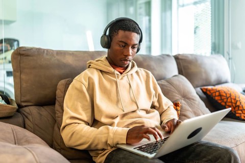 Young black man using laptop and headphones while working or studying from home, sitting on a