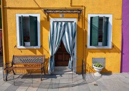 Yellow house facade with entrance door with green and white curtain and windows, colourful houses