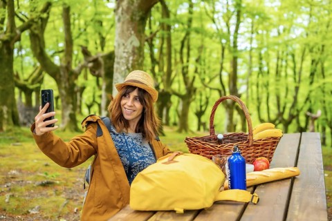 A young Caucasian woman waving on the phone sitting at a picnic table in the woods. Summer