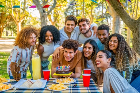 Young man smiling about to blow candles celebrating birthday with multi-ethnic friends at park