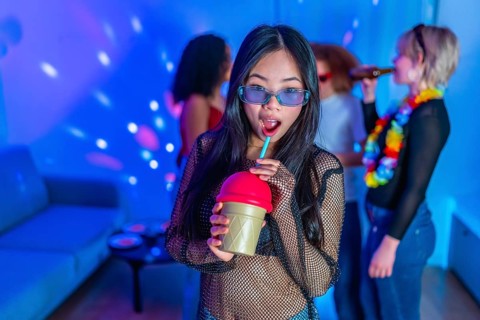 Asian woman drinking a colorful beverage at a lively party with friends, enjoying the music and