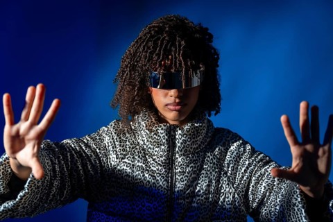 Young woman with futuristic visor and patterned jacket pushing virtual screen on blue background