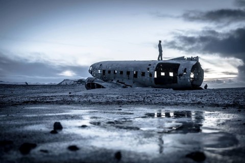 A man stands on the side of a crashed airplane. The sky is cloudy and the sun is setting