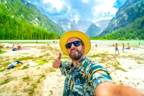 Tourist wearing sunglasses and straw hat gesturing thumbs up while taking a selfie at lago di