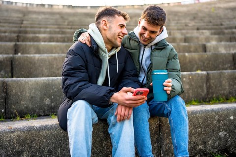 Two men are sitting on steps, enjoying a reusable coffee cup and sharing a laugh as they look at a