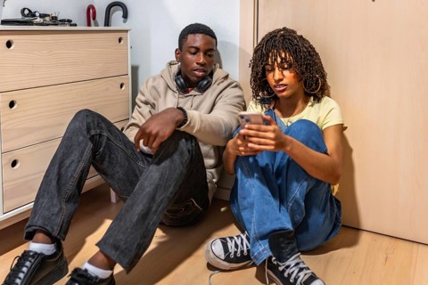 Two teenagers sitting on the floor of a cozy bedroom, using a smartphone and enjoying moments of
