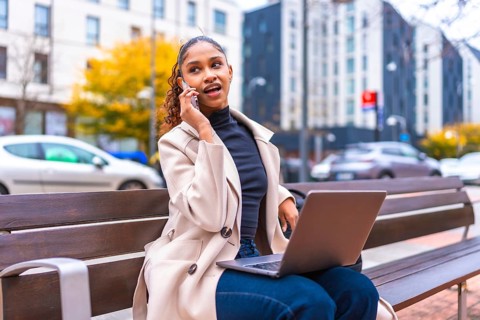 Busy young african american businesswoman using laptop talking to phone sitting on bench in the