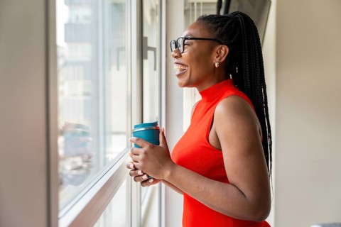 Happy businesswoman enjoys a coffee break in her hotel room, looking out the window and smiling