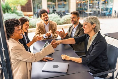 Businesswomen closing deal shaking hands in a cafeteria while colleagues applauding